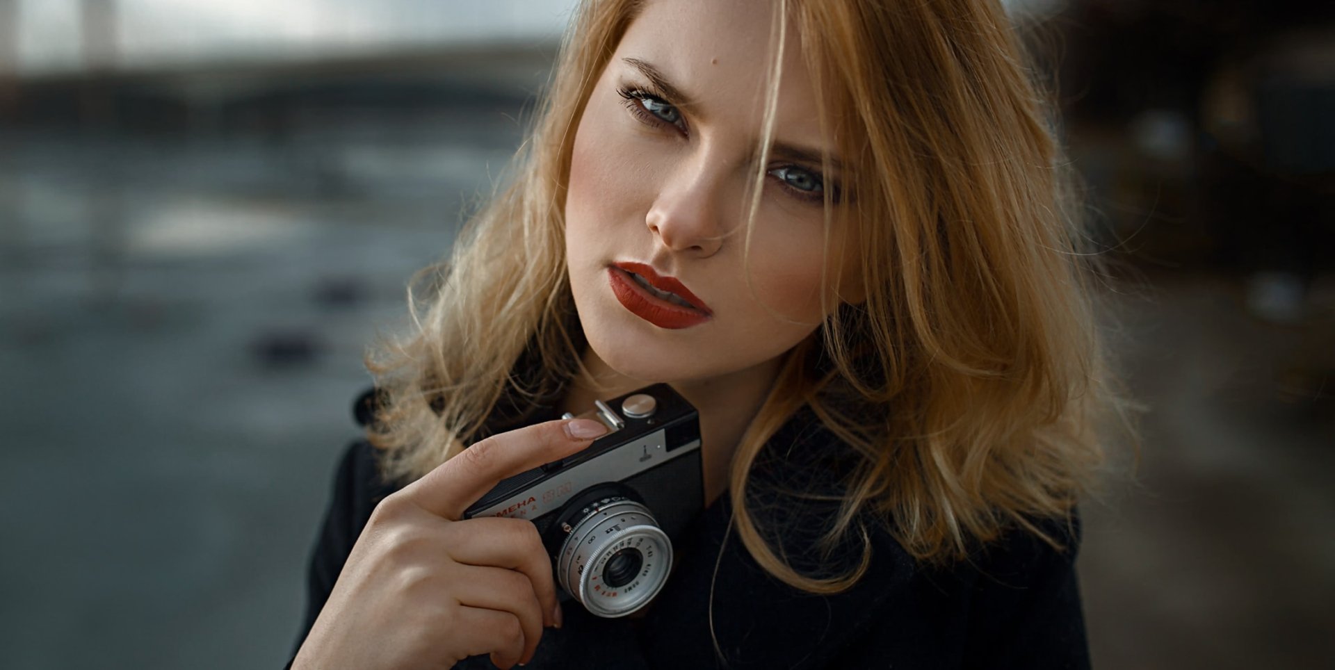 HD PC desktop wallpaper: close-up of a blonde female model with blue eyes and red lipstick holding a vintage camera; shallow depth of field softly blurs the background.