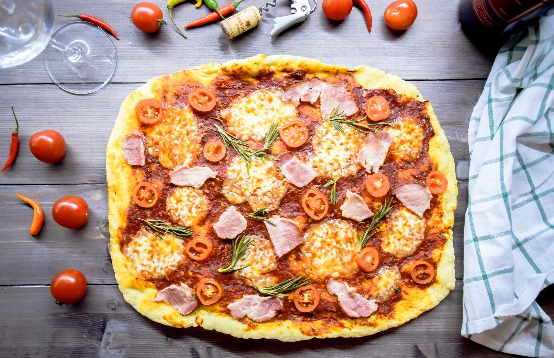 HD PC desktop wallpaper: food still life — overhead of a rustic tomato pizza topped with mozzarella, cherry tomatoes, ham and rosemary on a wooden table with scattered peppers.