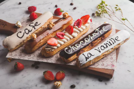 HD desktop wallpaper featuring a still life of four éclairs with café, chocolate, and vanilla flavors, adorned with fresh strawberries and nuts on a rustic wooden board.
