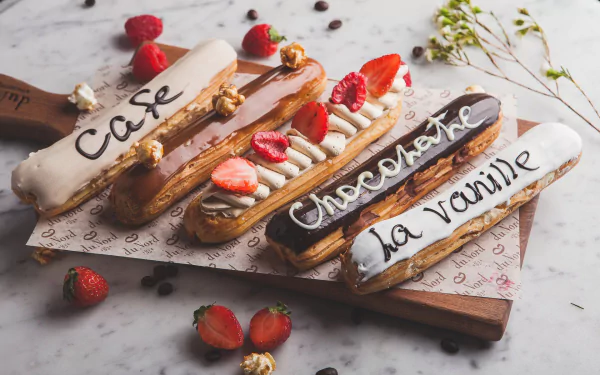 HD desktop wallpaper featuring a still life of four éclairs with café, chocolate, and vanilla flavors, adorned with fresh strawberries and nuts on a rustic wooden board.