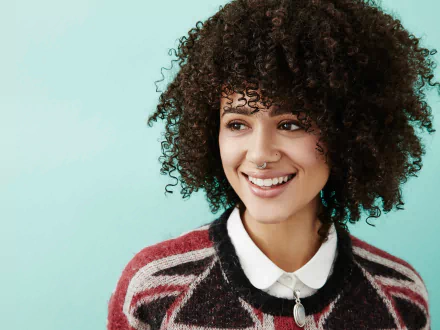 4K Ultra HD desktop wallpaper: smiling brunette woman with curly hair, brown eyes and a nose piercing, wearing a collared sweater against a teal background.
