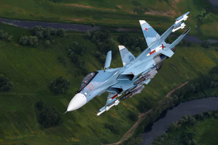 A Sukhoi Su-30 jet fighter soars through the sky, showcasing its military prowess against a backdrop of lush green landscapes and a winding river. HD desktop wallpaper for aviation enthusiasts.