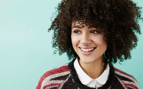 4K Ultra HD desktop wallpaper: smiling brunette woman with curly hair, brown eyes and a nose piercing, wearing a collared sweater against a teal background.