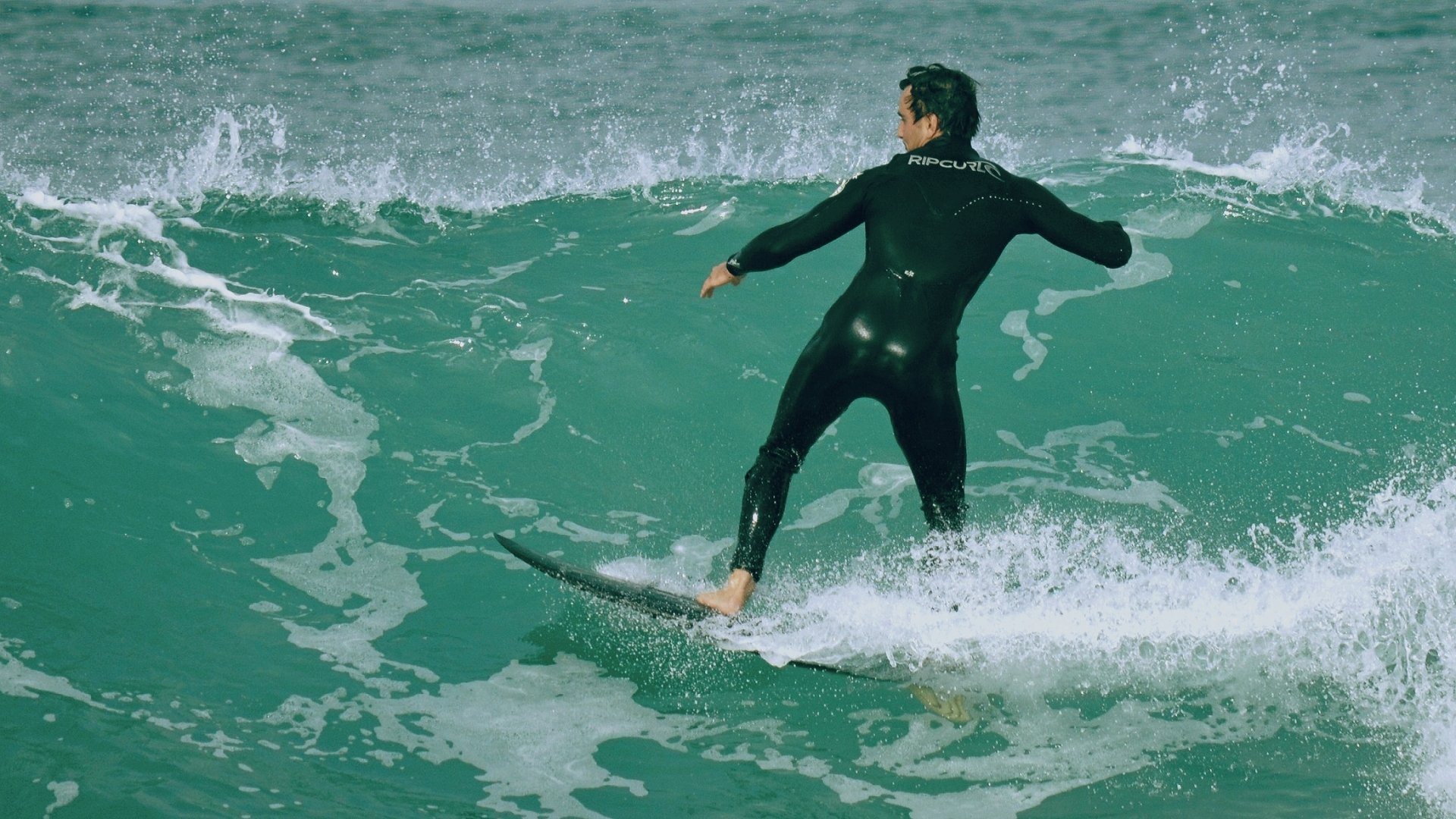 Surfer in a wetsuit riding green ocean waves — surfing sports action captured as an HD PC desktop wallpaper/background.