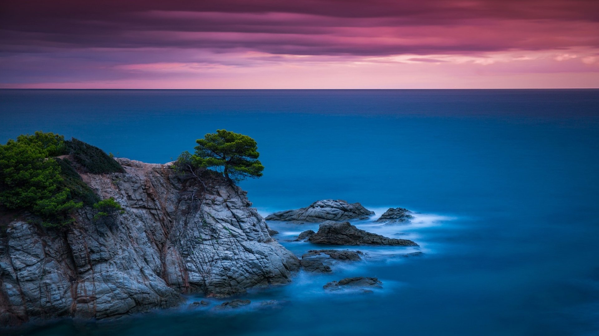 HD desktop wallpaper of a serene ocean horizon at dusk. A rocky shoreline with a lone tree overlooks the calm blue waters under a colorful, cloudy sky. Nature's beauty captured vividly.