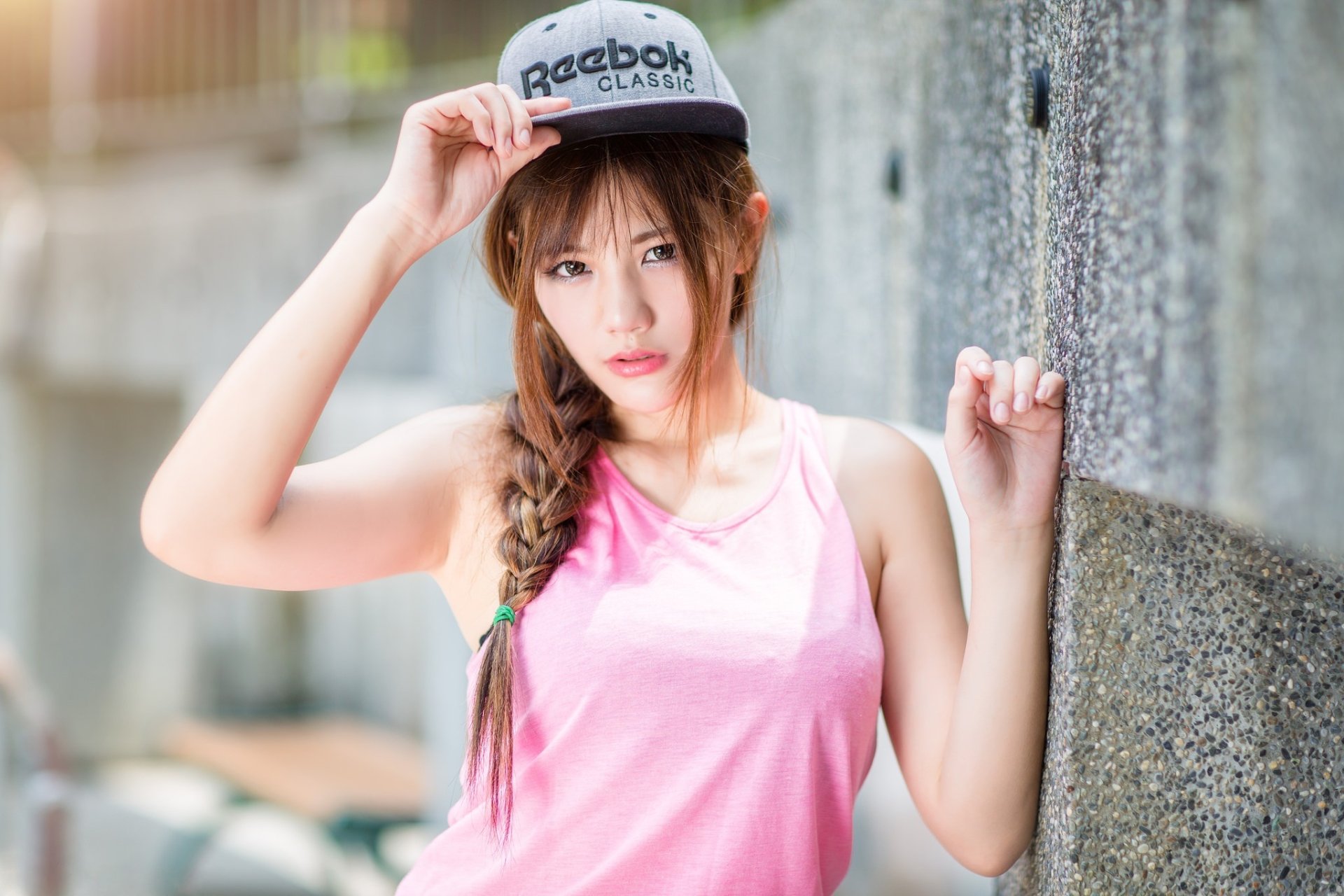 HD wallpaper of an Asian brunette woman with a braided hairstyle wearing a Reebok cap and a pink tank top, posing against a textured wall with a blurred background.