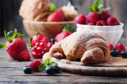 HD desktop wallpaper showing a breakfast scene with a croissant on a wooden board, surrounded by strawberries, raspberries, and blueberries. A bowl of raspberries and another croissant are in the background.