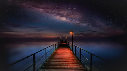 A HD wallpaper featuring a starry sky and the Milky Way galaxy over a serene ocean. A man-made pier stretches out into the night, illuminated by warm lights against the backdrop.