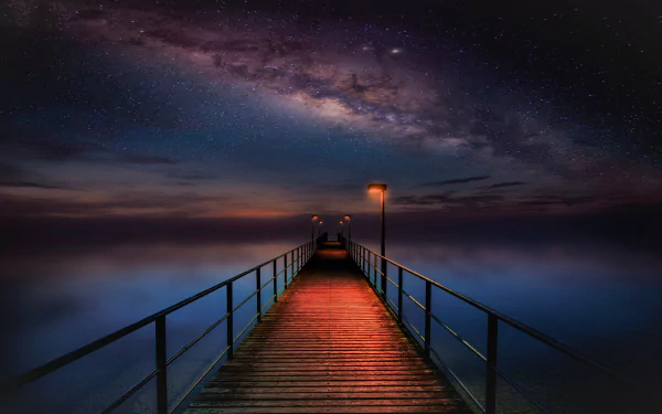 A HD wallpaper featuring a starry sky and the Milky Way galaxy over a serene ocean. A man-made pier stretches out into the night, illuminated by warm lights against the backdrop.