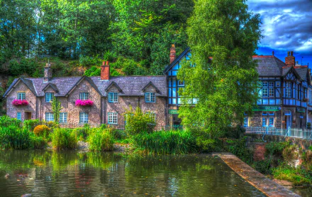 HDR photograph of a picturesque brick house in England, with window boxes full of flowers, a large tree, and a tranquil pond, capturing the essence of a serene countryside setting.