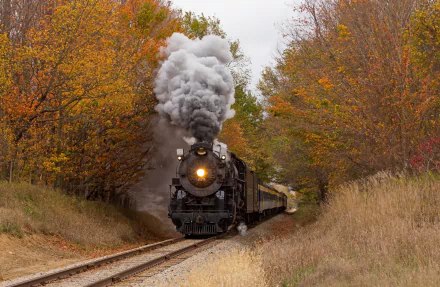A steam train chugs along a railroad surrounded by vibrant fall foliage, exuding smoke from its locomotive. This HD desktop wallpaper captures the nostalgic essence of classic rail travel amidst autumn scenery.