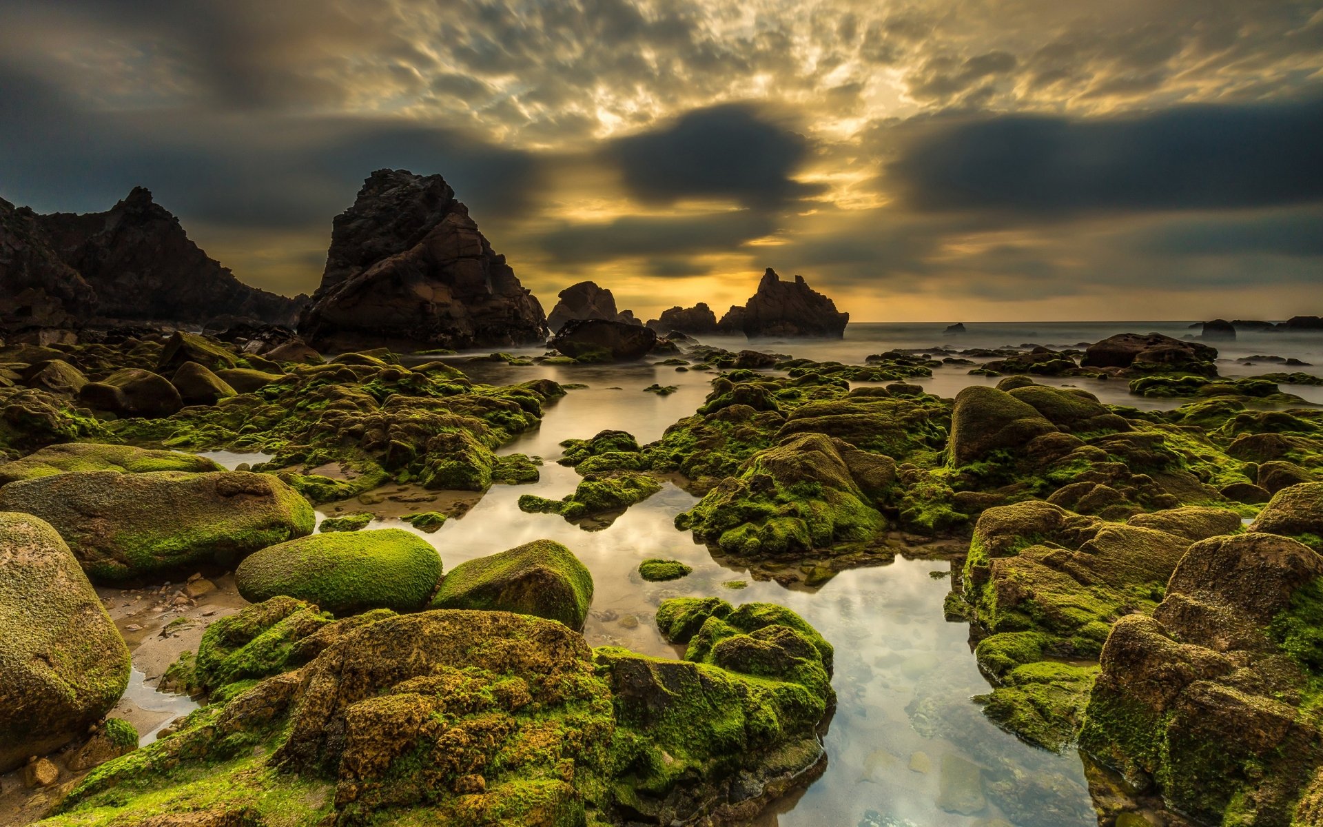 HD desktop wallpaper of a moss-covered rocky shoreline at sunset in Portugal, showcasing a dramatic seascape beneath a cloudy horizon.