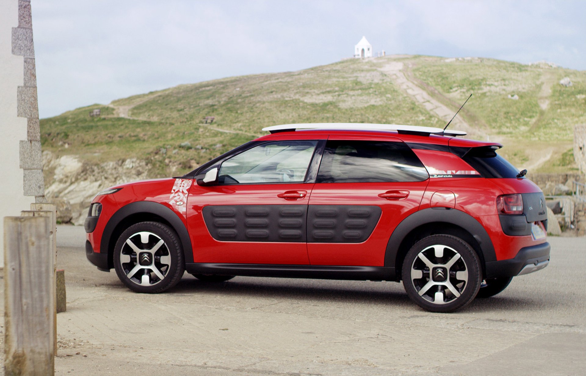 HD desktop wallpaper featuring a red Citroën C4 Cactus SUV parked on a beach with a grassy hill and small white building in the background.
