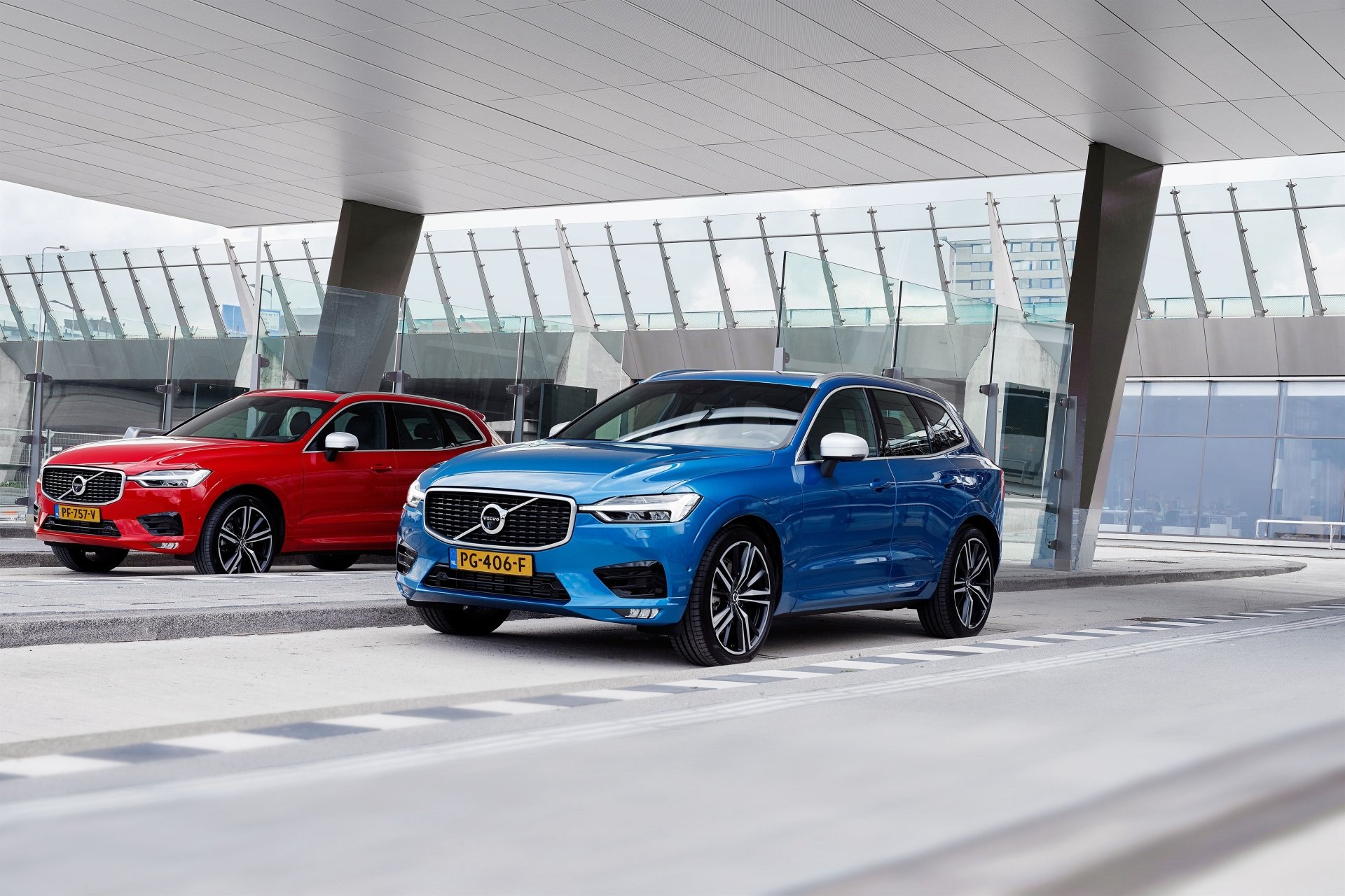 Blue Volvo XC60 SUV with a red Volvo behind it, parked under a modern glass canopy — 4K Ultra HD PC desktop wallpaper showcasing Volvo vehicles.
