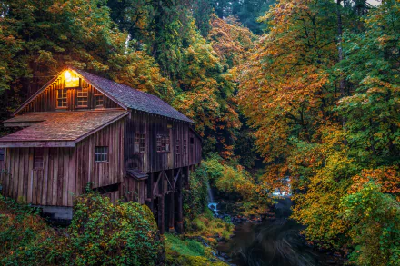 HD desktop wallpaper of a rustic watermill surrounded by vibrant autumn foliage in a serene forest setting. The illuminated window casts a warm glow over the scenic landscape.