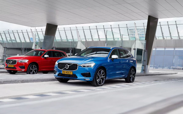 Blue Volvo XC60 SUV with a red Volvo behind it, parked under a modern glass canopy — 4K Ultra HD PC desktop wallpaper showcasing Volvo vehicles.