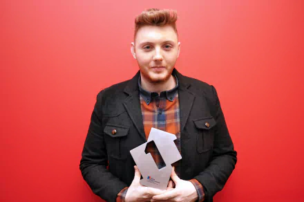 HD desktop wallpaper featuring a man with styled hair and a black jacket, against a red background, holding an award.