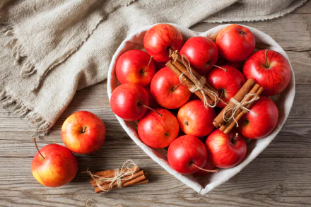 A heart-shaped bowl filled with red apples and tied cinnamon sticks on a wooden table, captured in a vibrant 4K Ultra HD still life food wallpaper.