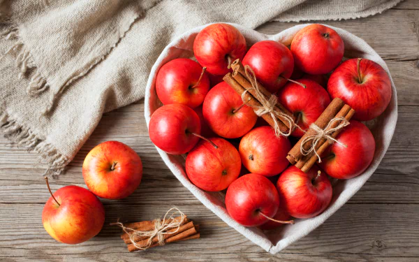 A heart-shaped bowl filled with red apples and tied cinnamon sticks on a wooden table, captured in a vibrant 4K Ultra HD still life food wallpaper.