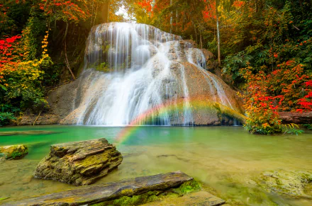 A vibrant 4K Ultra HD image of a serene lagoon with a cascading waterfall surrounded by autumn foliage and a vivid rainbow arching over the water.