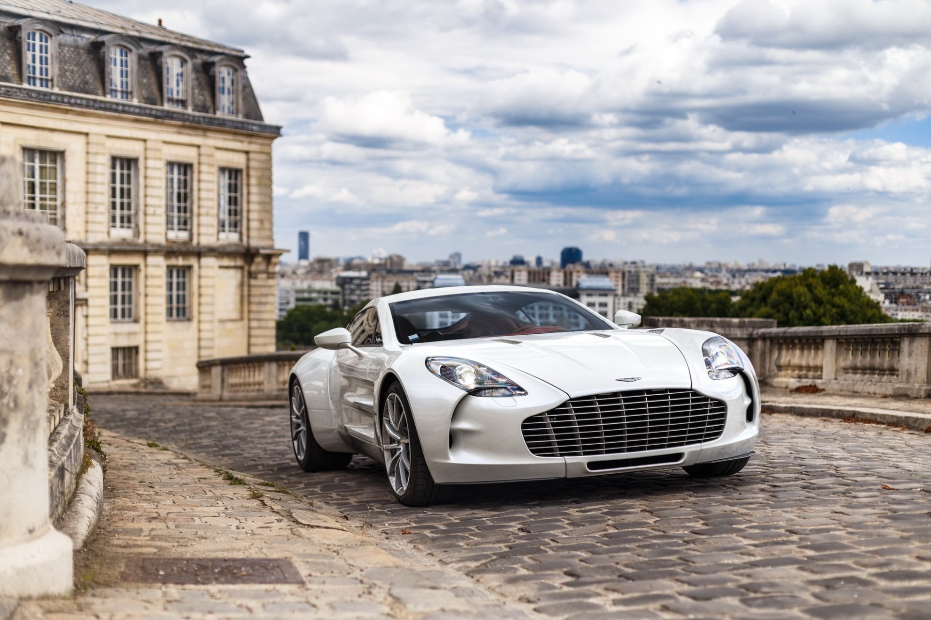 A white Aston Martin One-77 supercar parked on a cobblestone street with historic buildings and a cityscape under a cloudy sky in the background.