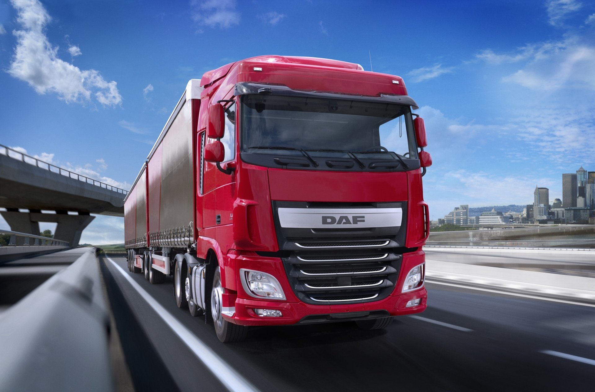Red DAF vehicle (semi-truck) speeding on a highway with city skyline under blue sky — 2K Quad HD PC desktop wallpaper/background.