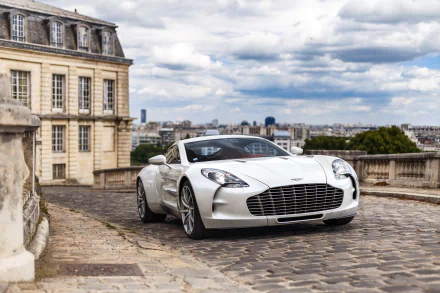 A white Aston Martin One-77 supercar parked on a cobblestone street with historic buildings and a cityscape under a cloudy sky in the background.