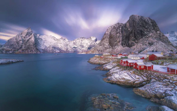 Snow-covered mountains rise over the tranquil lake in Reine, Lofoten, Norway, with red houses dotting the winter town under a moody sky.