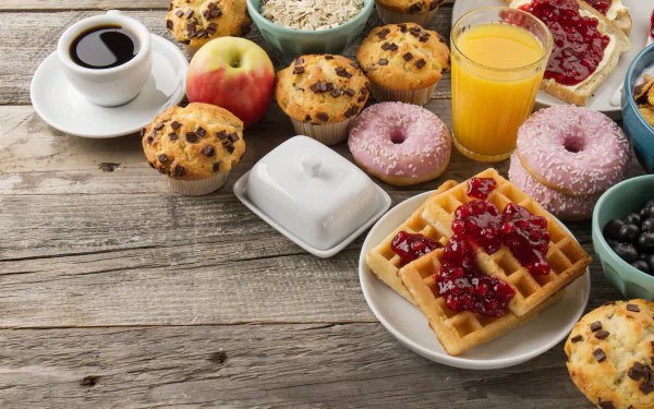 A vibrant 4K Ultra HD desktop wallpaper featuring a breakfast spread with muffins, waffles topped with jam, doughnuts, a cup of coffee, orange juice, and fresh fruit on a rustic wooden table.
