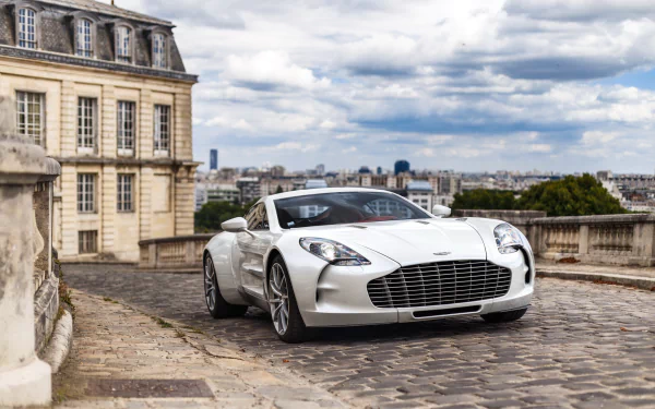 A white Aston Martin One-77 supercar parked on a cobblestone street with historic buildings and a cityscape under a cloudy sky in the background.
