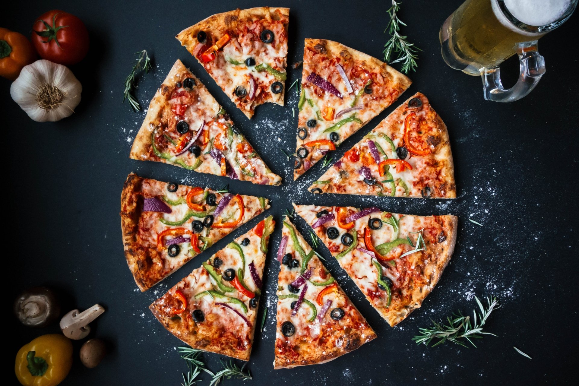 Top-down view of sliced pizza topped with capsicum, pepper, and olives, accompanied by a glass of beer, garlic, tomatoes, and herbs on a dark surface in 4K Ultra HD.