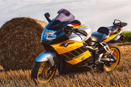 A BMW K1200S motorcycle with yellow and black accents is parked beside a hay bale in a field under a cloudy sky, captured in 4K Ultra HD.