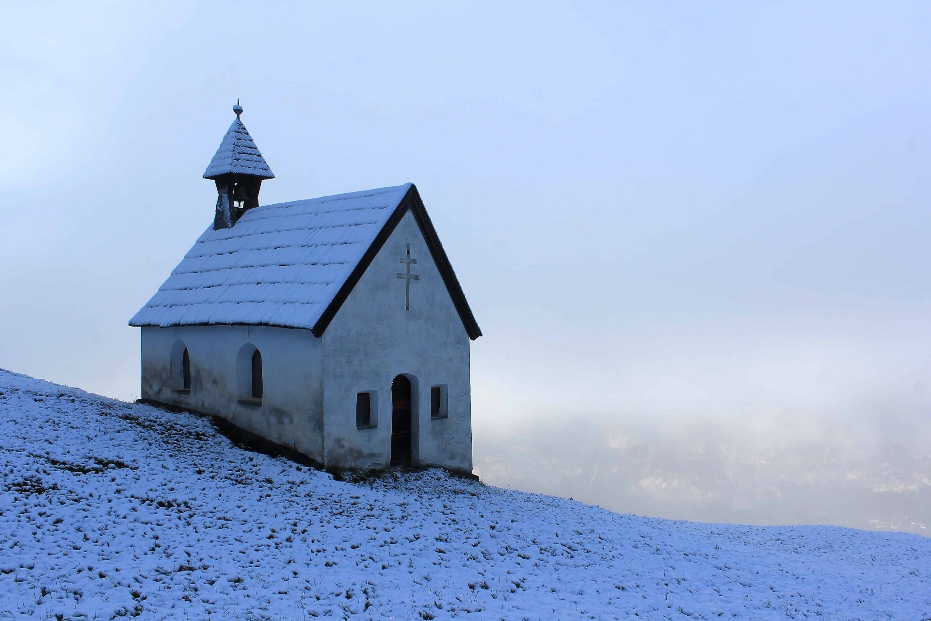 4K Ultra HD PC desktop wallpaper and background: a small snow-covered chapel on a winter hillside, a solitary religious church silhouette beneath a pale, misty sky.