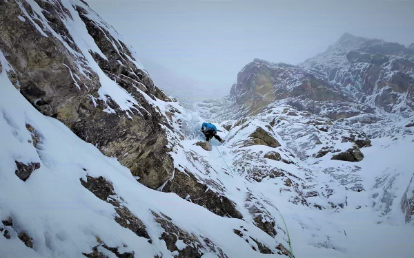 A climber in winter gear ascends a steep, snowy mountain slope amidst rugged rocky terrain, captured in a detailed 4K Ultra HD mountaineering scene.