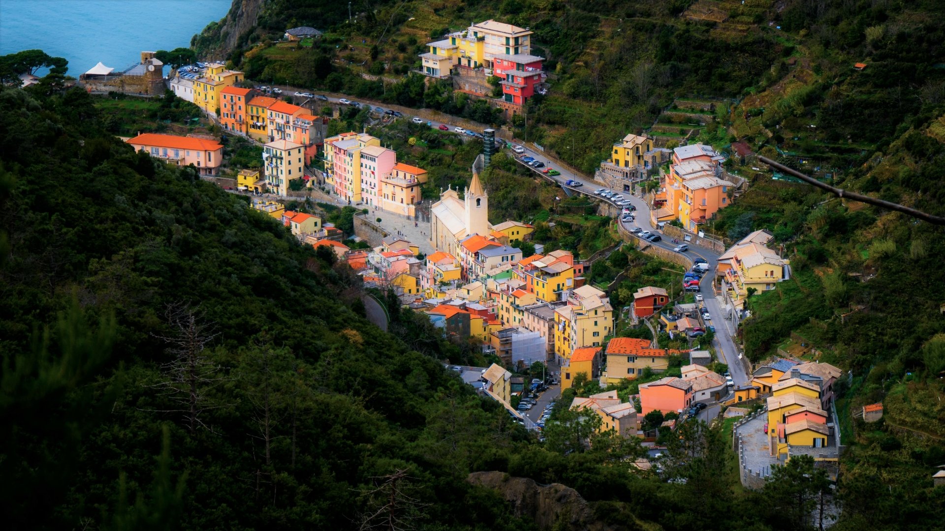 Colorful Ligurian village of pastel houses and a central church, nestled in terraced green hills by the sea — 4K Ultra HD PC desktop wallpaper of a man-made hillside settlement.