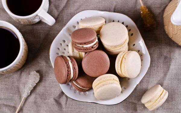 A 4K Ultra HD desktop wallpaper featuring a plate of assorted macarons alongside cups of coffee on a textured fabric surface, highlighting sweets and food themes.