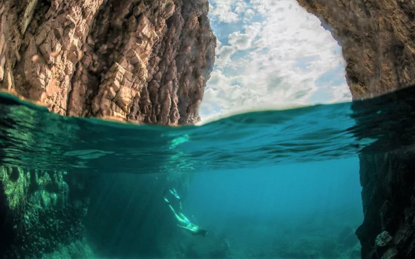 A scuba diver explores a stunning underwater cave, framed by striking rock formations and illuminated by sunlight peeking from above, creating a captivating ocean scene.
