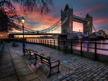 HD desktop wallpaper of Tower Bridge in London, England, at sunset. A serene street with benches lines the riverside, casting a tranquil scene against a colorful sky.