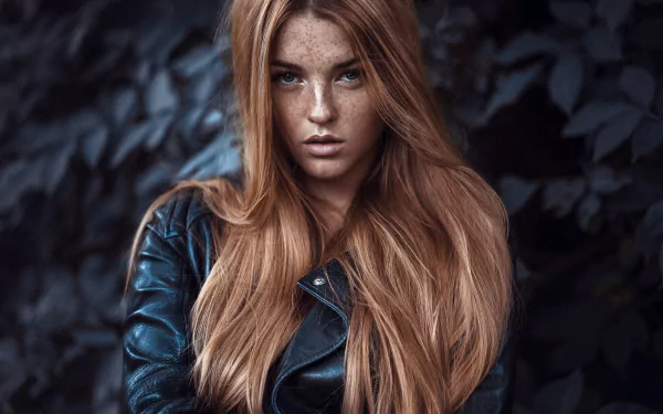 HD desktop wallpaper of a freckled redhead woman model with long flowing hair in a leather jacket against a moody, leafy background.