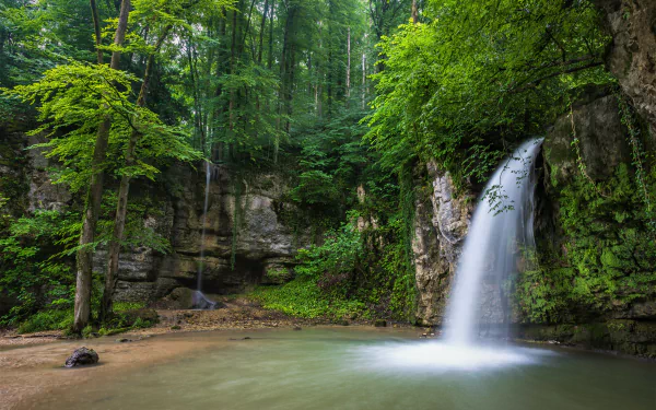 A serene HD desktop wallpaper featuring a lush green forest with a cascading waterfall flowing into a tranquil pool below. The scene captures the beauty and tranquility of nature.