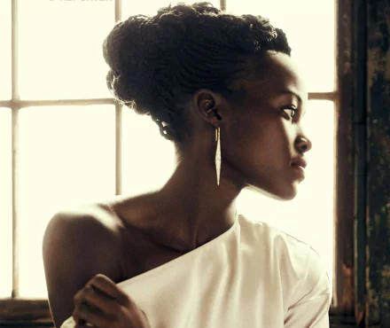 HD PC desktop wallpaper: profile portrait of a celebrity woman with braided updo and gold earring, wearing a white one-shoulder top, bathed in soft window light and warm tones.