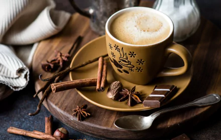 A warm cup of coffee surrounded by star anise, cinnamon sticks, chocolate pieces, and whole nutmeg, arranged as a cozy still life on a wooden surface.
