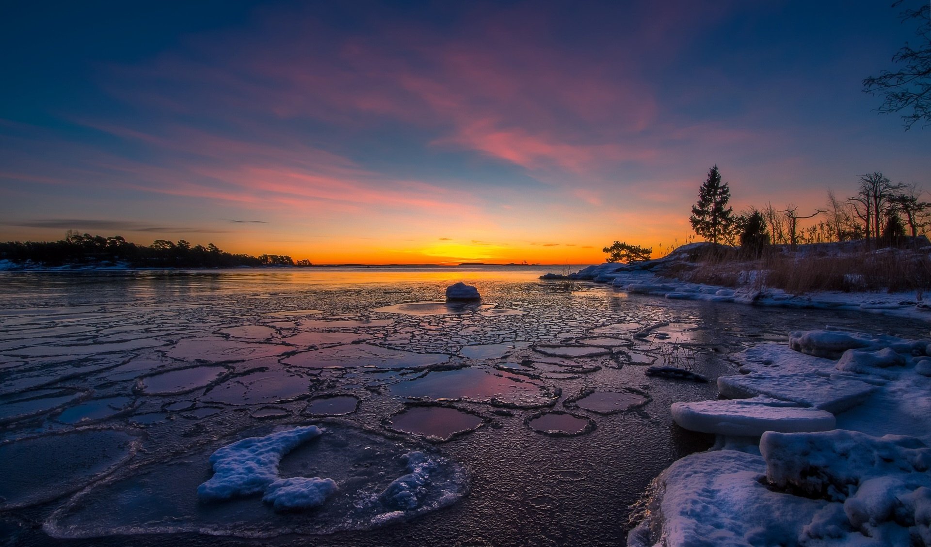 A stunning HD winter desktop wallpaper featuring a frozen lake with cracked ice at sunrise, under a vibrant sky, surrounded by nature.