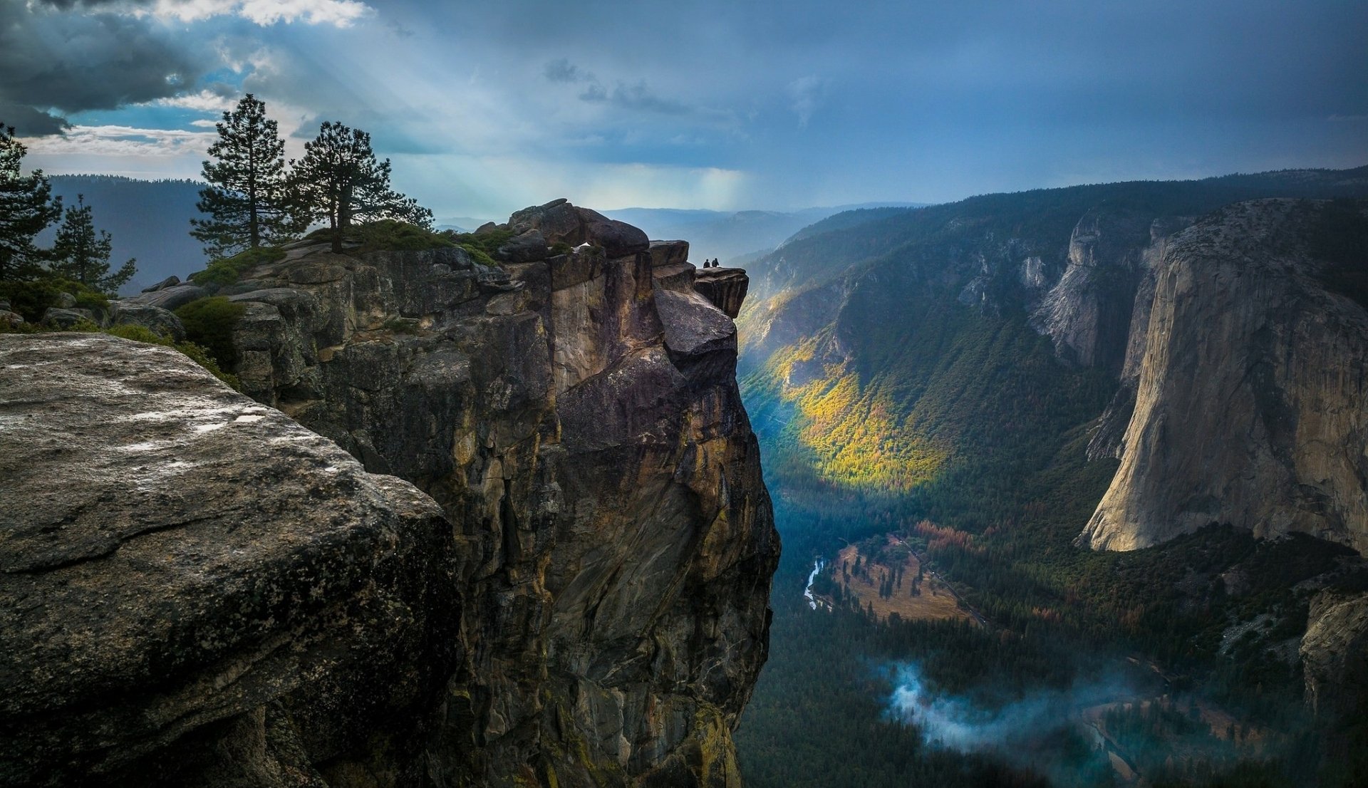 HD desktop wallpaper of a stunning landscape featuring a forested valley and rugged mountains, with dramatic cliffs and rays of sunlight piercing through the clouds.