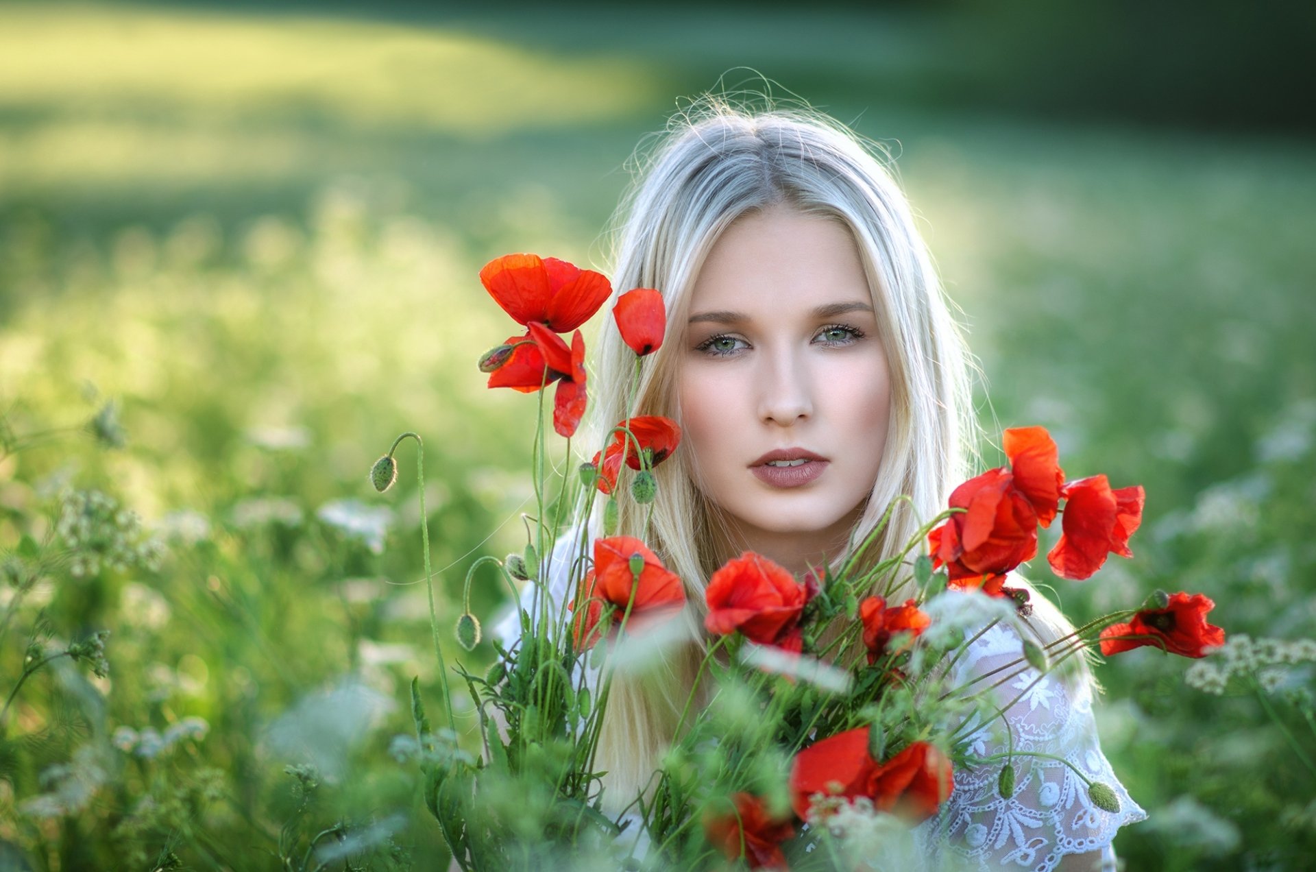 Blonde woman with green eyes surrounded by vibrant red poppy flowers in a sunlit summer field, captured in sharp focus with a soft depth of field background.