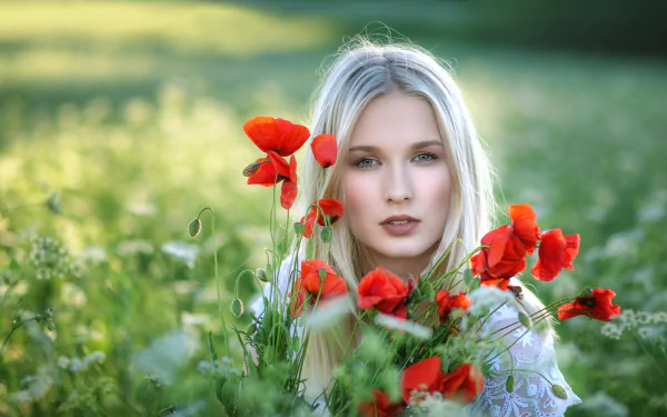 Blonde woman with green eyes surrounded by vibrant red poppy flowers in a sunlit summer field, captured in sharp focus with a soft depth of field background.
