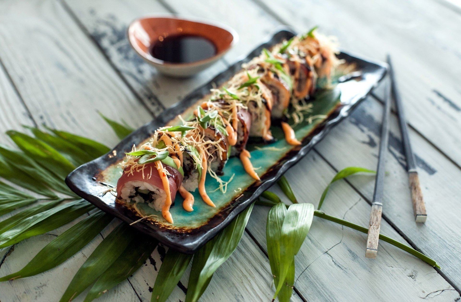 HD desktop wallpaper featuring a still life of sushi rolls with fish, rice, and sauce, accompanied by chopsticks and a dish of soy sauce on a rustic wooden surface.