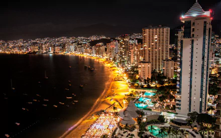 Nighttime view of Acapulco, Mexico, featuring illuminated city buildings and a beach along the coast. The HD wallpaper captures the vibrant nightlife and the stunning coastal scenery.