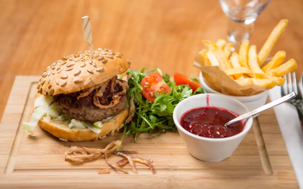 HD desktop wallpaper featuring a juicy burger with sesame bun, crispy french fries, fresh salad, and a cup of ketchup on a wooden serving board.