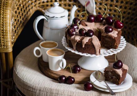 HD desktop wallpaper showing a chocolate cherry cake with frosting, two coffee cups, a teapot, and fresh cherries on a round table with a wicker chair.